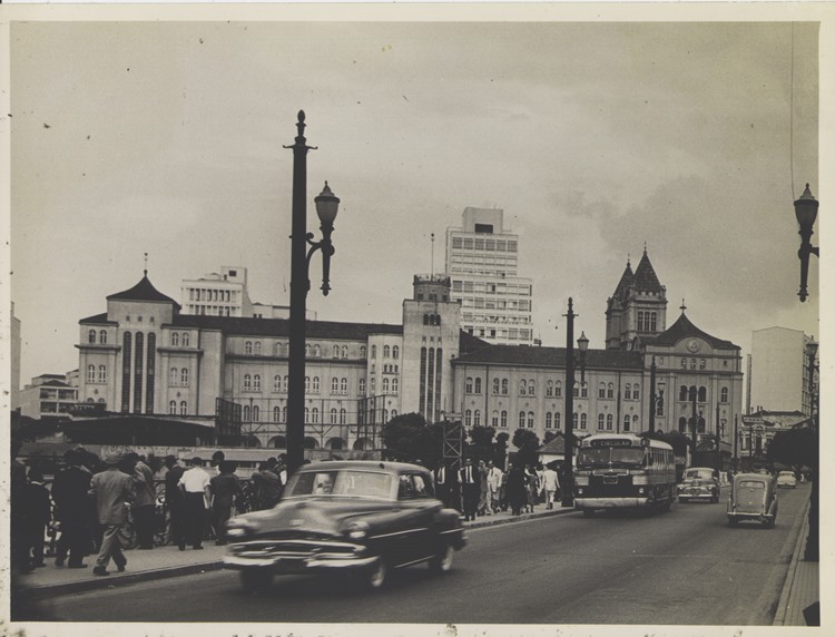 A imagem mostra o Viaduto Santa Ifigênia, em São Paulo, na década de 1950. Automóveis antigos, ônibus e pedestres movimentam-se em frente ao Mosteiro de São Bento, destacando arquitetura histórica e o dinamismo urbano da época.