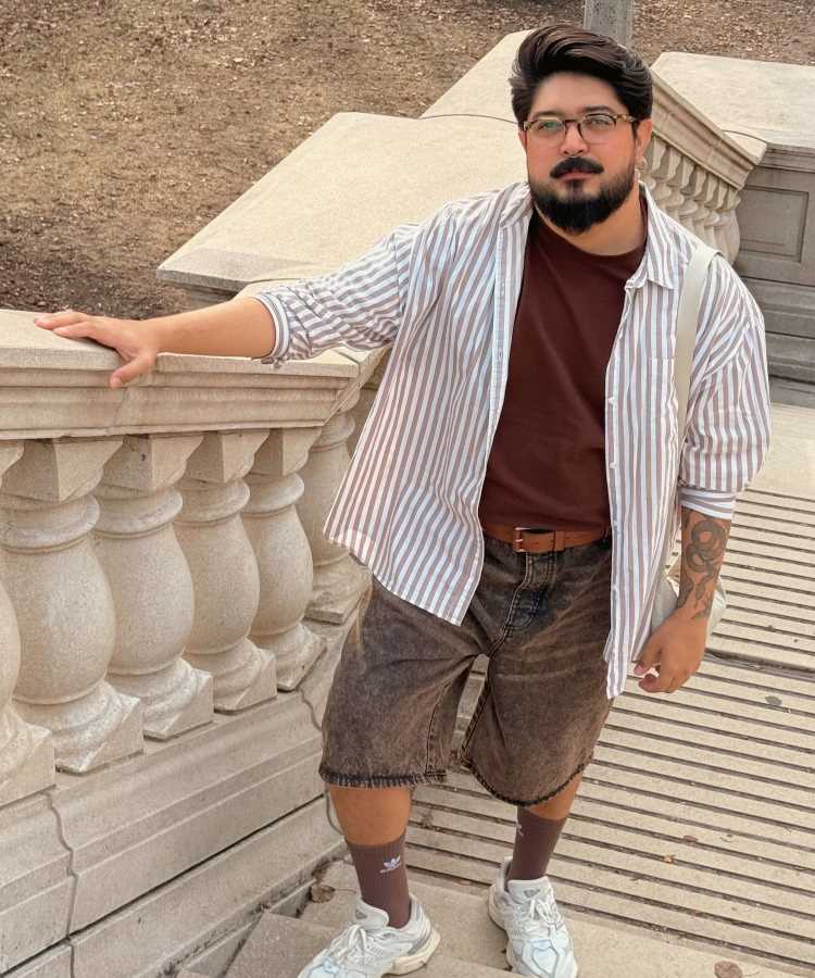 Homem com cabelos escuros, barba e óculos posa próximo a uma escadaria de pedra. Ele veste camiseta marrom, camisa listrada, bermuda jeans desfiada, meias escuras e tênis brancos, transmitindo um estilo casual e despojado em cenário urbano.