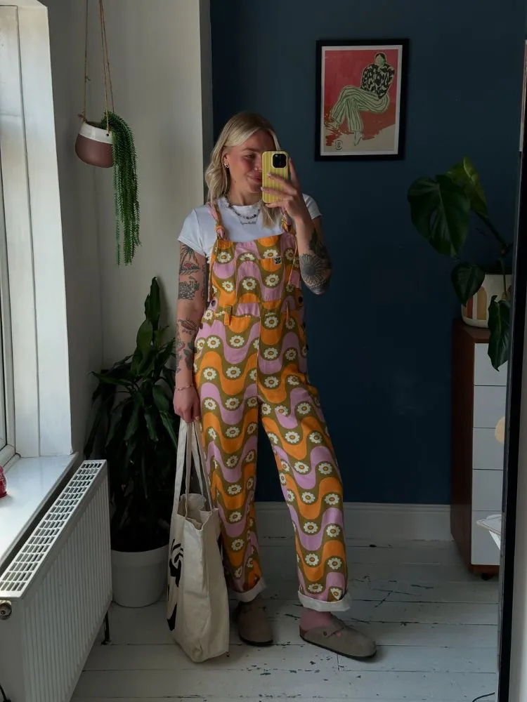 Mulher de pele clara usando blusa branca, jardineira floral em tons de verde, laranja e rosa, com clogs e ecobag