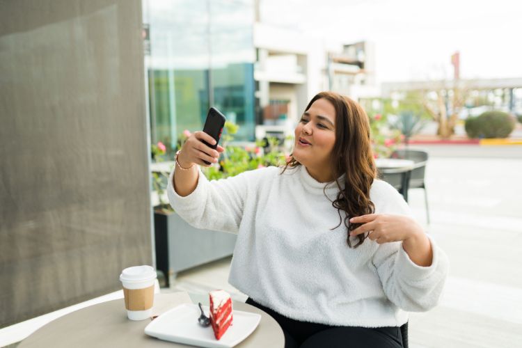 Mulher em café com bolo e café, fazendo selfie com expressão animada. A cena representa a criação de conteúdo e interação com seguidores, ilustrando o tema “diferença entre influencer e blogueira” no contexto de lifestyle digital e redes sociais.