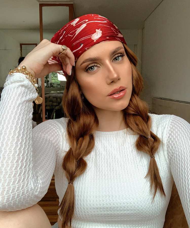 Mulher com cabelos castanhos e ondulados em duas tranças, usando lenço vermelho estampado e blusa branca texturizada, posando em ambiente interno.
