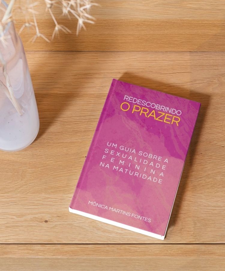 Livro "Redescobrindo o Prazer" de Mônica Martins Fontes sobre uma mesa de madeira. A capa é rosa, com título em branco e amarelo. Ao lado, um vaso branco com galhos decorativos, compondo um ambiente acolhedor.