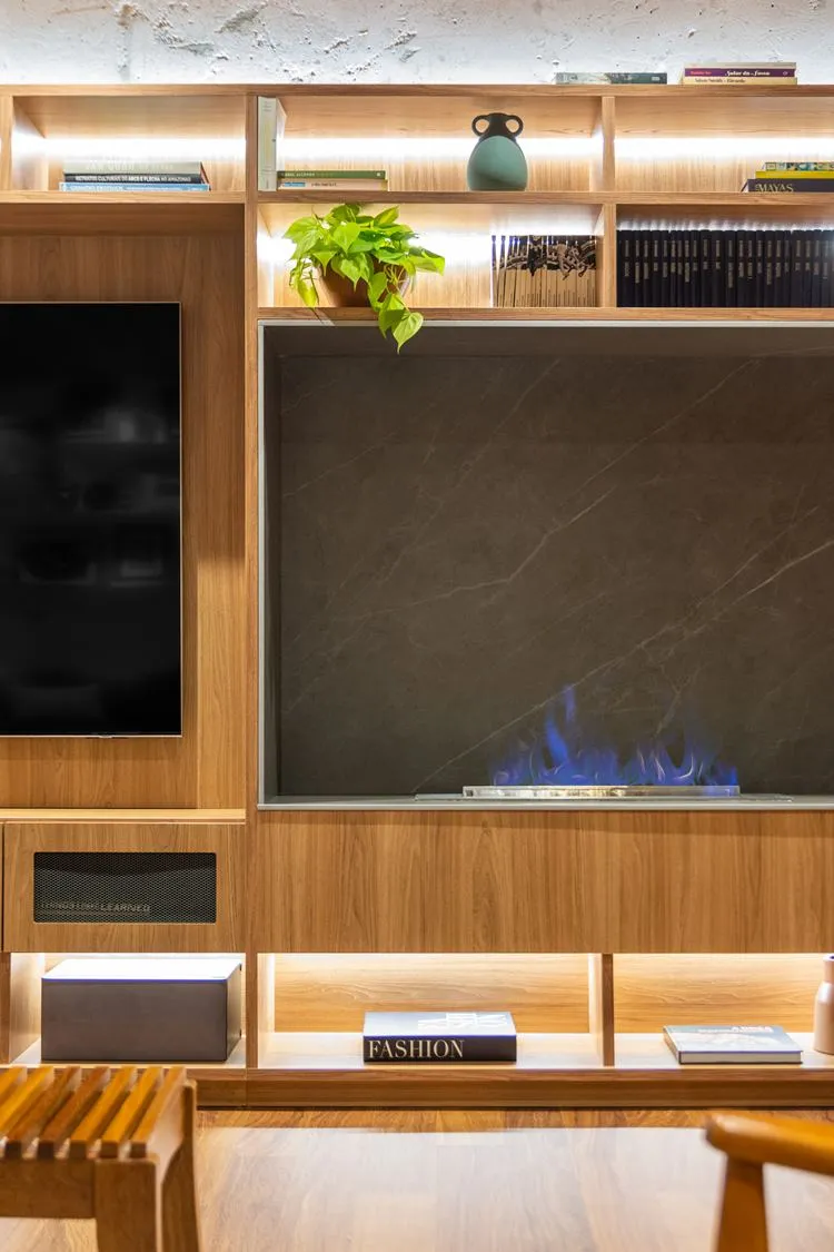 Lareira no home-theater do apartamento decorado de 187 m². Acabamento em porcelanato que imita mármore cinza. Planta jiboia, livros, caixas e outros objetos decoram as repartições do móvel