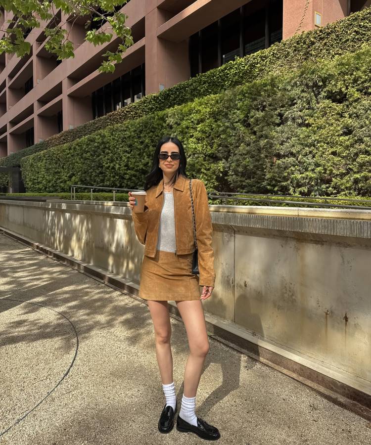 Mulher de corpo inteiro usando um conjunto de saia e jaqueta de camurça marrom, blusa cinza e mocassins pretos com meias brancas. Ela está de óculos de sol e segura um copo de café enquanto posa em um ambiente urbano com edifícios e vegetação ao fundo.








