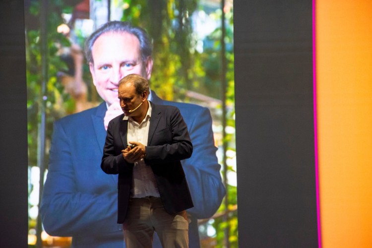 Alex Allard palestrando usando terno azul, camisa clara e calça bege com uma foto dele mesmo atrás no telão