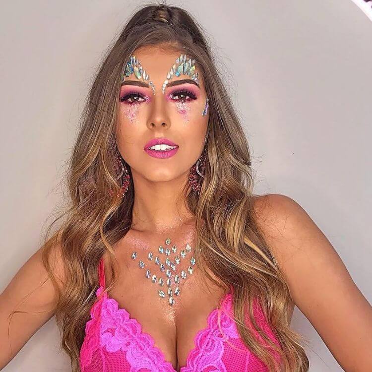 Mulher de pele clara usando maquiagem rosa, glitter abaixo dos olhos e pedrarias na testa e colo