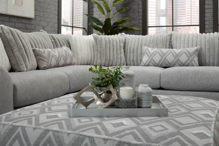 A imagem mostra uma sala moderna com sofá cinza em "L", decorado com almofadas texturizadas e geométricas em tons neutros. Sobre o puff central, há uma bandeja prateada com velas, vasos decorativos e uma planta. Grandes janelas iluminam o ambiente.