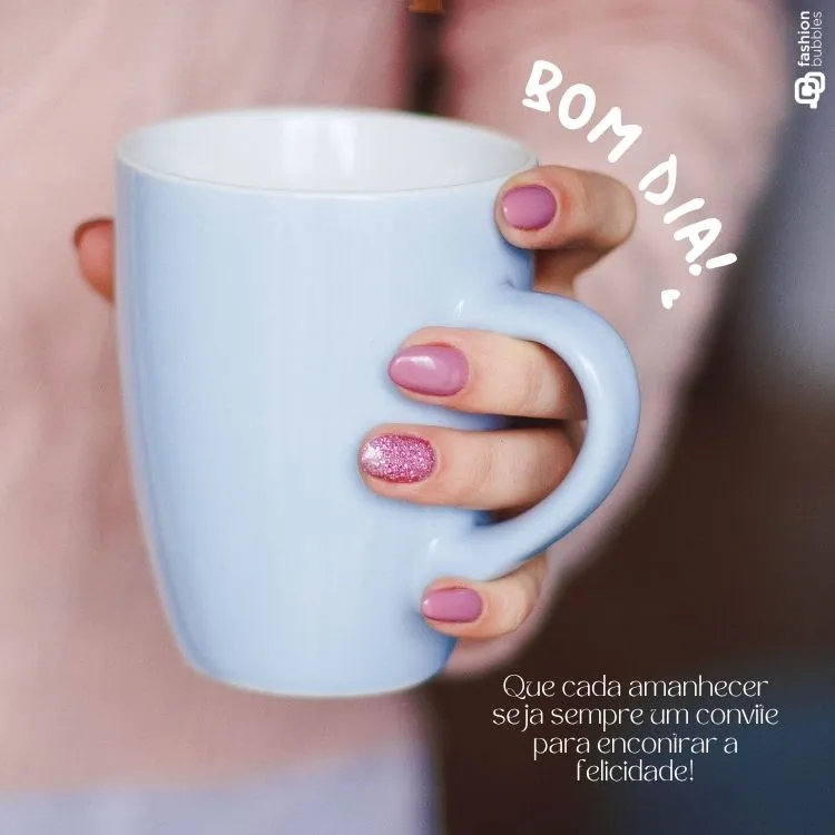 Uma mão feminina com unhas bem cuidadas, pintadas de rosa claro e uma com glitter, segura uma caneca azul-clara. A imagem evoca a sensação de um bom dia e convida à felicidade.