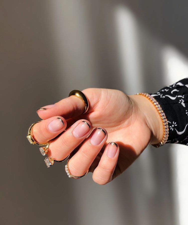 A imagem mostra uma mão com unhas decoradas em um estilo de francesinha moderna. As unhas têm uma base transparente com uma linha preta fina nas pontas. A mão está adornada com vários anéis dourados e um anel com uma pedra grande. O pulso tem uma pulseira brilhante. A luz na imagem destaca o brilho dos acessórios e o design das unhas.