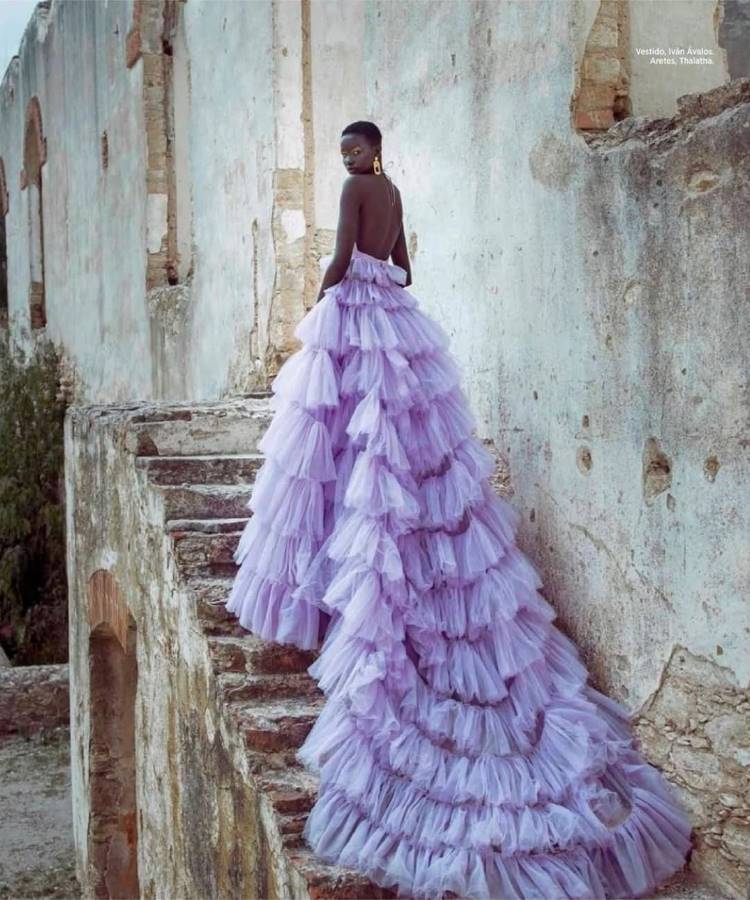 Uma modelo de pele escura com cabelo curto e brincos dourados, vestindo um volumoso vestido de tule lilás com babados e cauda longa, sobe uma escadaria de pedra desgastada em ruínas.







