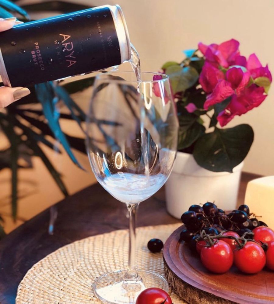 espumante em lata preta sendo despejado em taça de vidro. Ao lado, temos prato com tomates e uvas