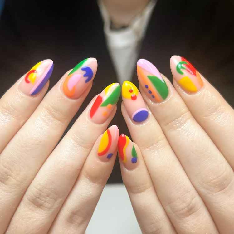 Unhas longas e ovais decoradas com esmalte em tons vibrantes de amarelo, laranja, verde, azul e vermelho, criando um design abstrato e colorido. O fundo escuro contrasta com as cores vivas, destacando o estilo ousado e artístico das unhas.