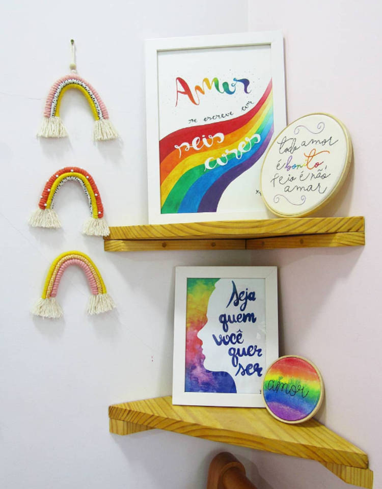 Cantinho com as cores da bandeira LGBTQIA+ em quadro escrito "amor se escreve em seis cores", bastidor com frase "todo amor é bonito, feio é não amar", quadro d emoldura branca com frase "seja quem você quer ser", bastidor colorido escrito "amor" e 3 arco-íris de macramê pendurados