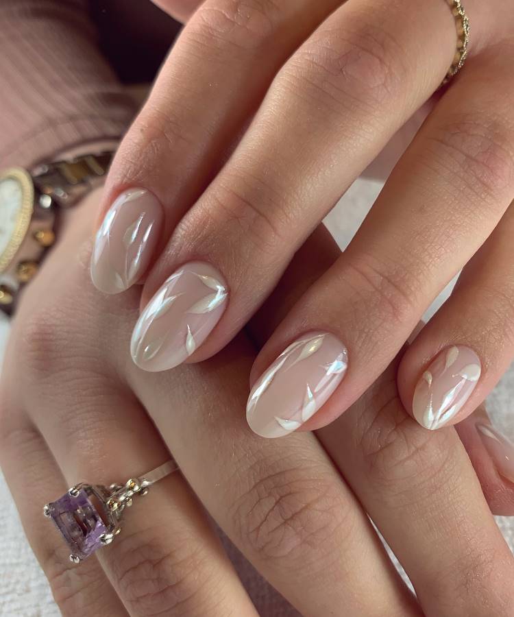 Mãos femininas com unhas em formato oval, decoradas com esmalte nude translúcido e delicados detalhes brancos com aparência de folhagens. Um anel dourado com pedra lilás e acessórios sutis complementam o estilo elegante. Fundo desfocado destaca o design requintado.






