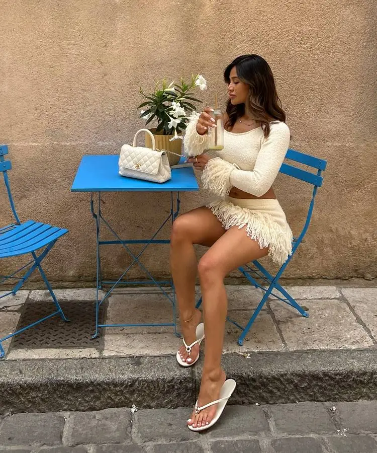 Uma mulher está sentada em uma mesa de café azul, bebendo uma bebida verde. Ela usa um conjunto de duas peças creme com franjas, uma pequena bolsa branca acolchoada e chinelos brancos de salto.