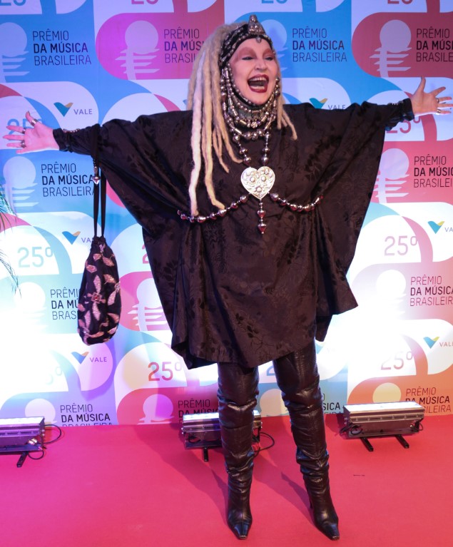 Foto colorida de Elke Maravilha mais velha posando sorrindo no 25º Prêmio da Música Brasileira em pé e de braços abertos, vestida toda de preto, com botas de cano alto, peruca loira estilo dreadlocks e muitos acessórios. Elke Maravilha posando sorrindo no 25º Prêmio da Música Brasileira em pé e de braços abertos, vestida toda de preto, com botas de cano alto, peruca loira estilo dreadlocks e muitos acessórios.