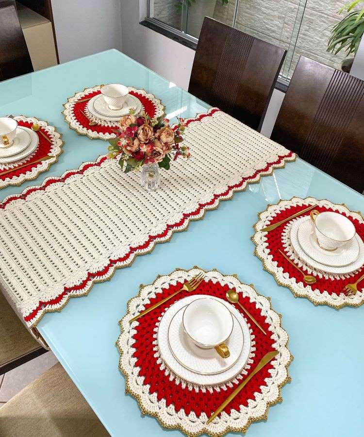 Mesa decorada com sousplats de crochê vermelho e dourado, louças brancas, talheres dourados. Centro de mesa com arranjo floral em vaso de vidro sobre toalha de crochê combinando.