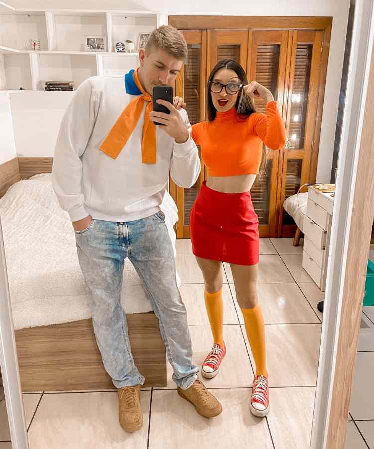 Fantasia de casal para o Halloween: Velma e Fred de Scoody Doo. Ele usa moletom branco com lenço laranja e calça jeans. Ela usa cropped laranja e saia vermelha 