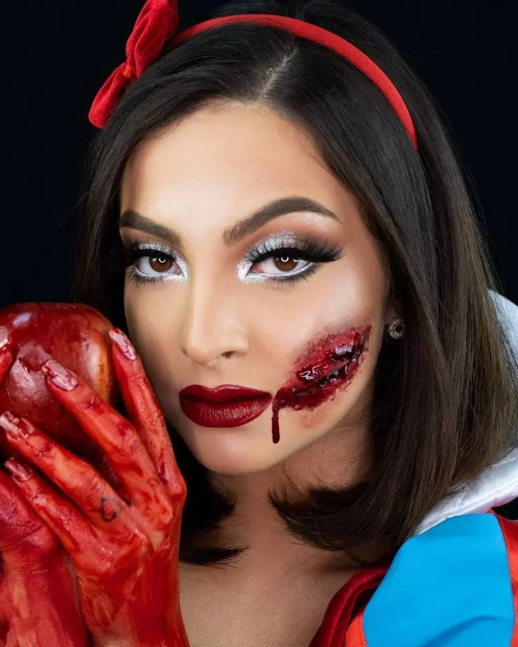 Mulher de pele clara usando fantasia de Branca de Neve com machucado de sangue falso e mão ensanguentada segurando maçã