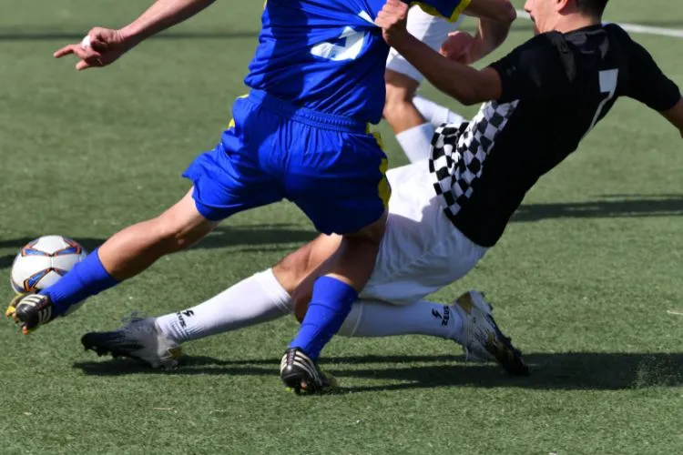 Três pessoas jogando futebol, uma ao fundo de bermuda branca e duas a frente disputando bola, uma de uniforme azul e outra de preto e branco 