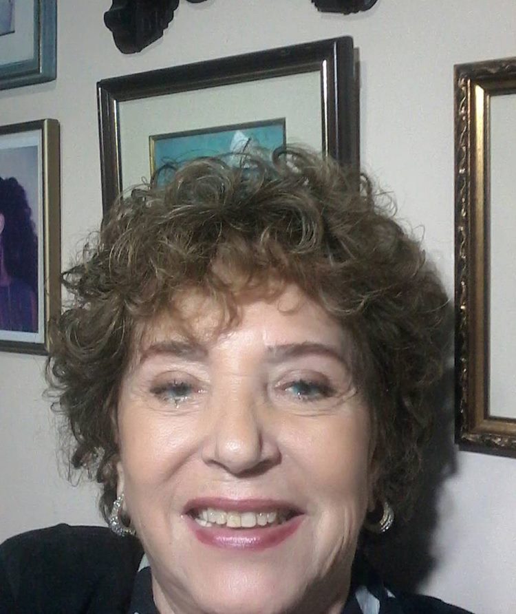 Um retrato de uma mulher de idade com cabelos cacheados castanhos, sorrindo gentilmente para a câmera. Ela usa brincos e uma blusa preta estampada. O fundo mostra uma parede com quadros decorativos.