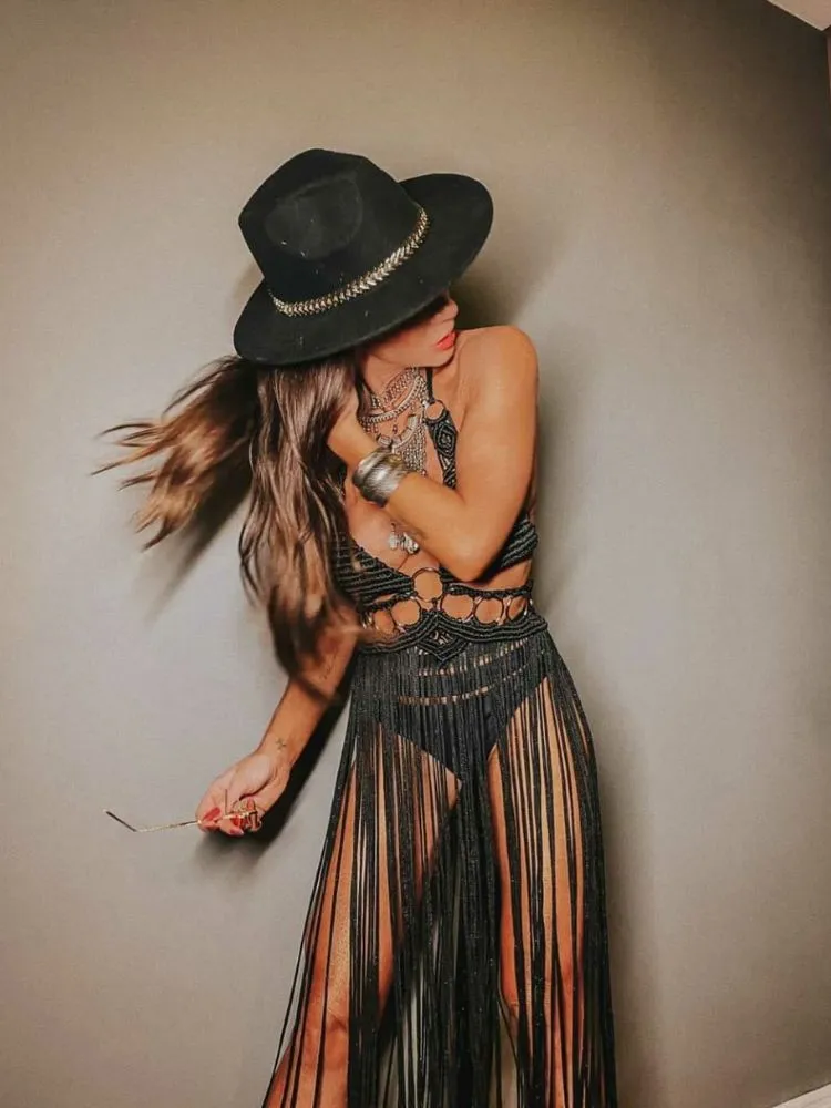 Mulher de pele clara usando chapéu preto, hot pant preta e vestido preto em macramê com transparência, franjas e ferragens