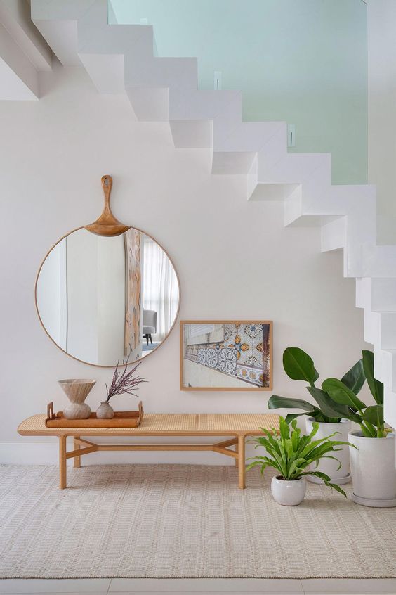 Hall de entrada branco com escada em cima, espelho, quadro, banco de madeira, bandeja decorativa e vasos brancos com planta sno chão 