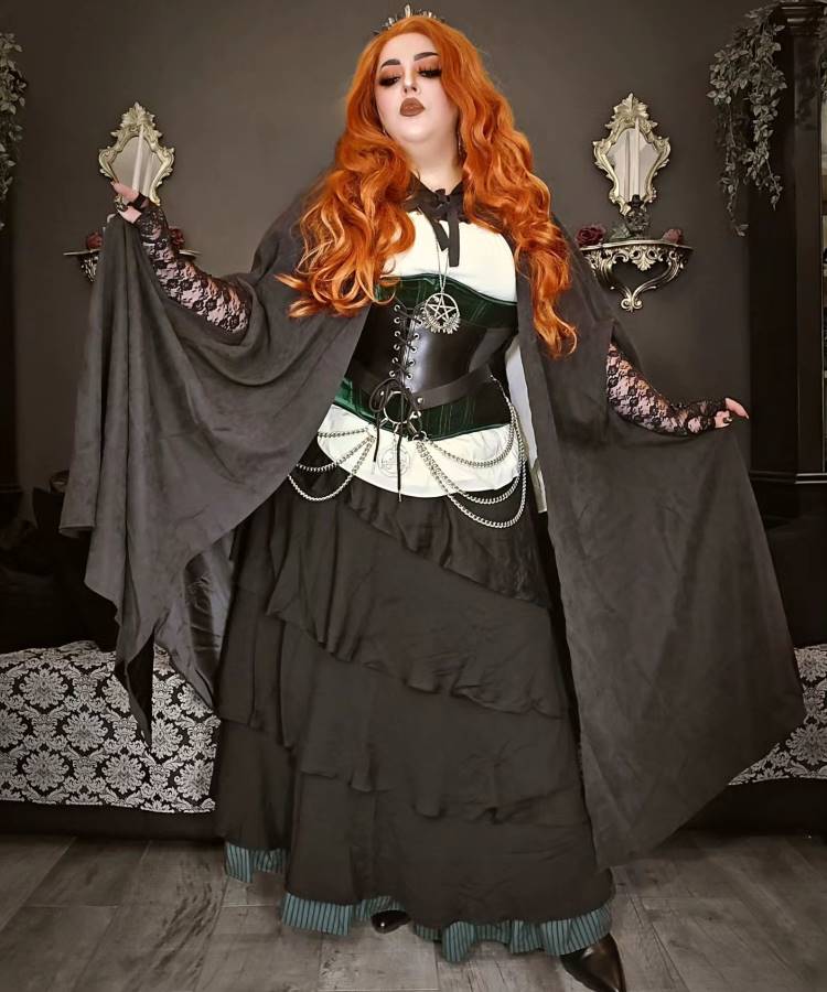 A imagem apresenta uma mulher com longos cabelos ruivos, usando uma fantasia gótica. Ela veste uma saia preta volumosa, corpete com detalhes em verde e correntes prateadas. As mangas de renda preta e uma capa escura completam o visual. O cenário é elegante, com decoração clássica, destacando as capas de Halloween como um elemento central do traje.


