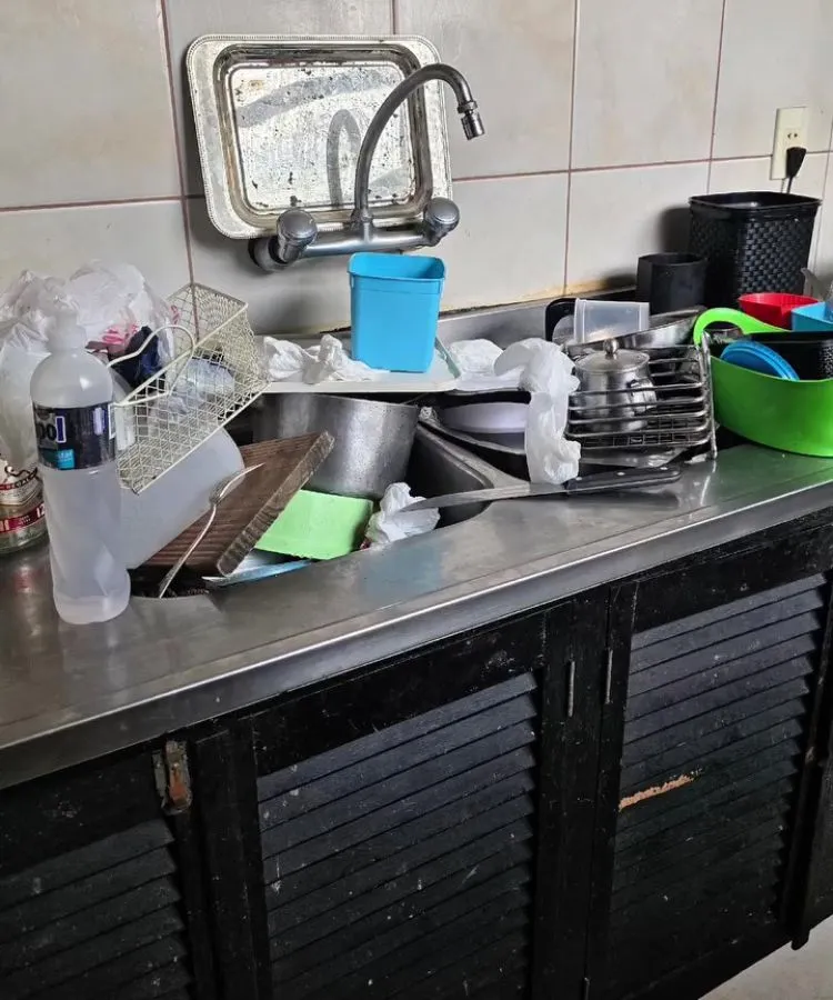 Pia de cozinha com louça suja acumulada, incluindo pratos, copos, talheres e panelas. Há uma garrafa plástica de álcool, esponja verde e recipiente azul sobre a bancada. Armário preto e torneira prateada completam o ambiente desorganizado, transmitindo descuido doméstico.