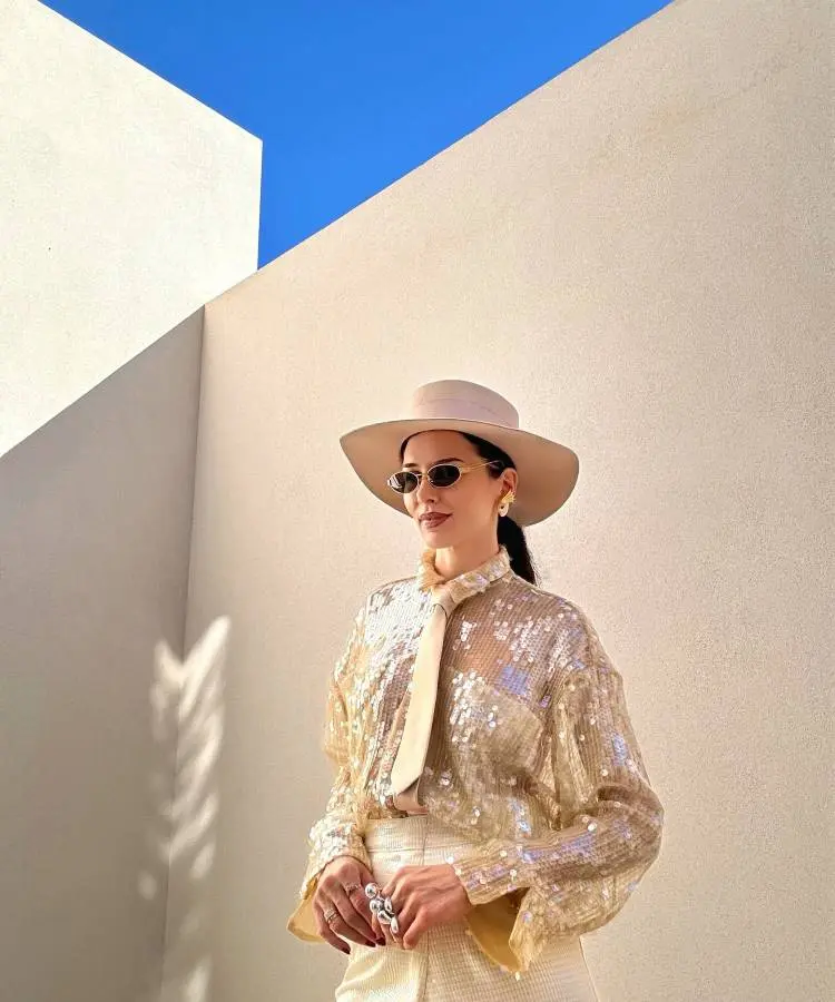 A mulher posa em um ambiente minimalista ao ar livre, vestindo uma blusa dourada de paetês, gravata bege e chapéu elegante. Os óculos escuros e o céu azul intenso complementam o visual sofisticado e moderno, em tons claros e brilhantes.