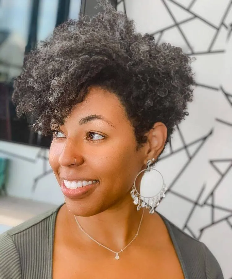 Uma mulher sorri para o lado, exibindo um corte de cabelo cacheado volumoso com tons grisalhos. Ela usa um brinco grande e circular, um piercing no nariz e um colar delicado sobre uma blusa escura.
