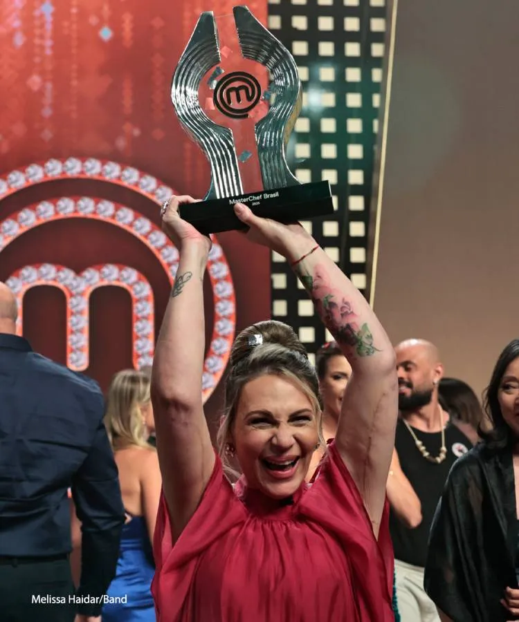 Uma mulher loira e sorridente, com uma blusa vermelha, levanta o troféu do MasterChef Brasil com os braços tatuados. Ela comemora sua vitória no palco, cercada por outras pessoas.