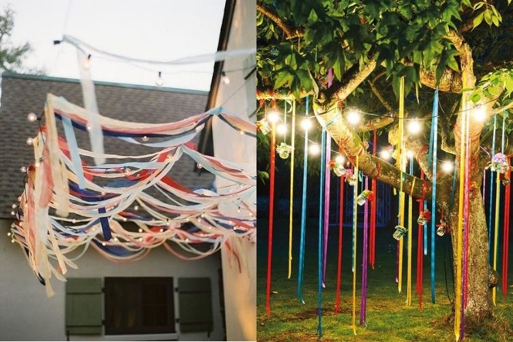 A imagem representa uma decoração de festa junina com firas. Contém fitas penduradas em árvore com luzes e fitas com luzes penduradas ao ar livre