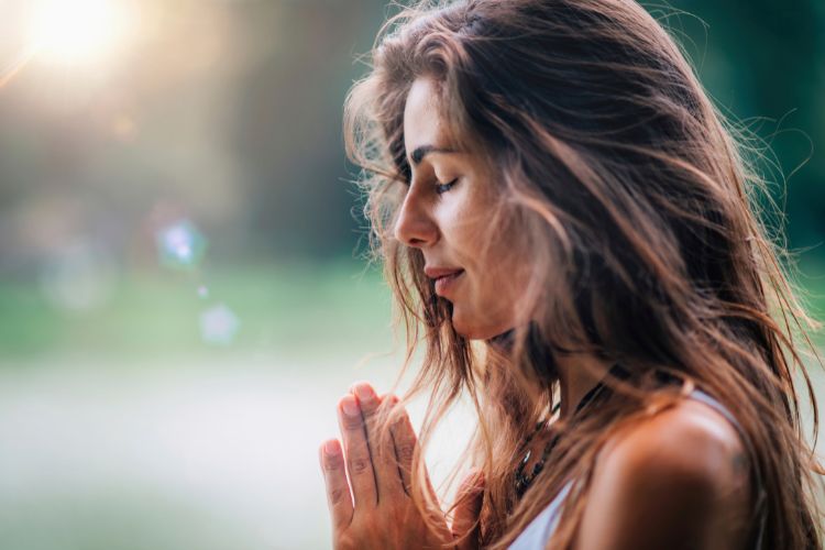 Mulher jovem de perfil, com cabelos ao vento, mãos em posição de prece, aparenta estar em meditação. Fundo desfocado com natureza e luz solar suave brilhando, transmite serenidade e conexão espiritual em ambiente ao ar livre.