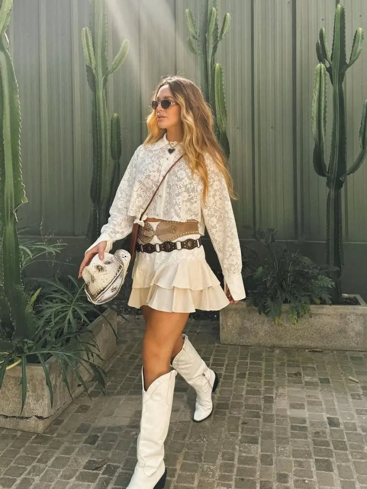 Mulher de pele clara de camisa de renda, saia de babados, dois maxi cintos, chapéu country e bota, tudo em branco 