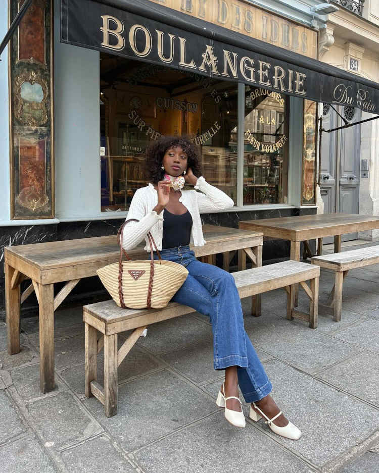 Mulher sentada em um banco, com uma blusa preta, um casaco branco e calças jeans. Nos pés estão um par de sapatos Mar Jane brancos com bico quadrado.