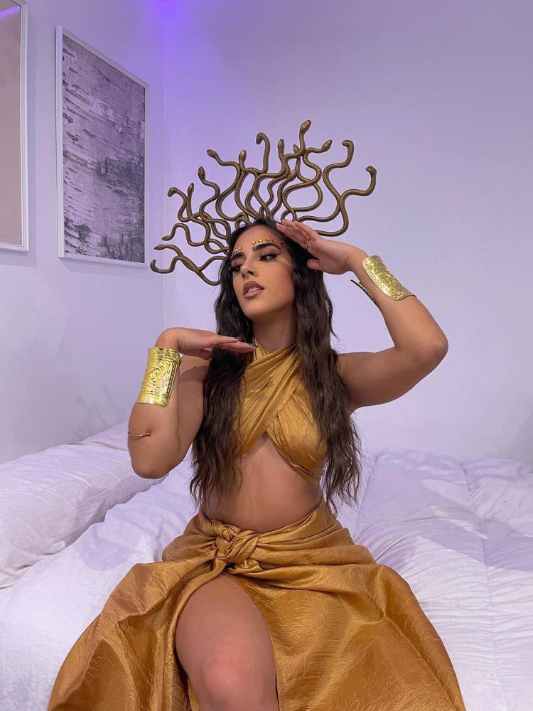 Mulher de pele clara sentada na cama usando tiara dourada de medusa e cropped e saia longa dourados 