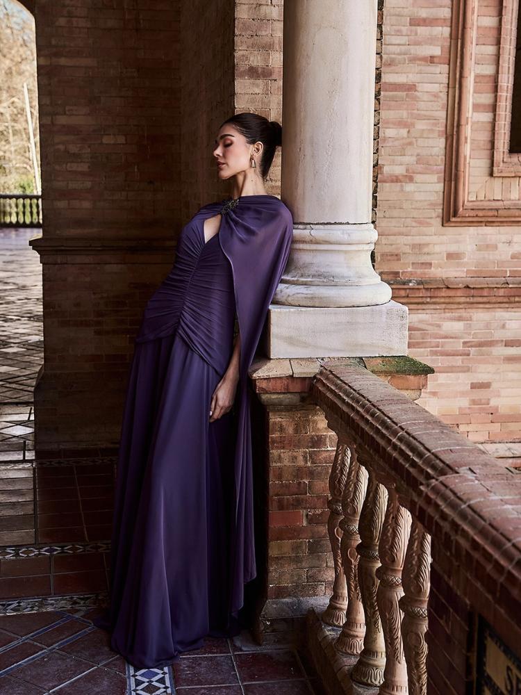 Mulher de pele clara com vestido roxo drapeado minimalista e capa com broche