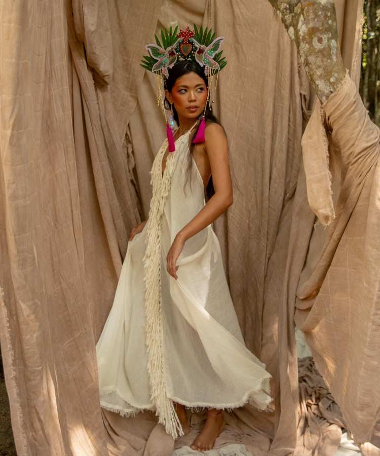 Mulher posa em meio a tecidos bege, usando vestido longo branco com franjas e adorno de cabeça colorido com folhas, pássaros e coração central. Brincos pink destacam-se. Ambiente natural com árvore ao fundo exala suavidade e elegância.