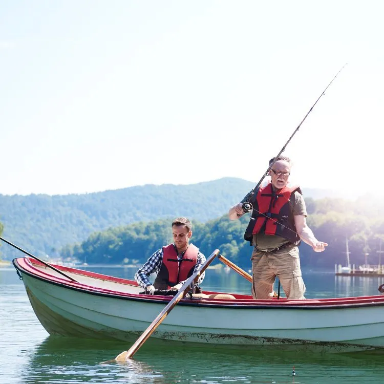 A imagem mostra duas pessoas em um barco, uma adulta e uma idosa, ambas com coletes salva-vidas. A pessoa idosa segura uma vara de pescar, sugerindo uma atividade de pesca. O cenário inclui água calma e montanhas ao fundo sob céu claro. É um momento de lazer que evoca o Dia dos Pais criativo.