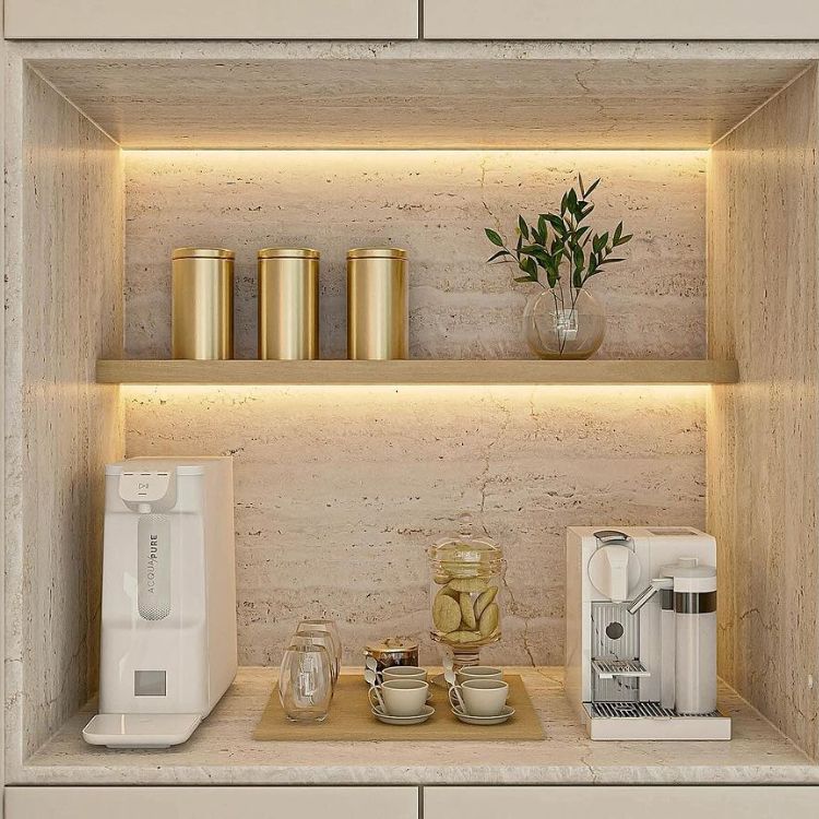Cantinho do café embutido em móvel bege com iluminação, cafeteria, xícaras, potes dourados e planta. 