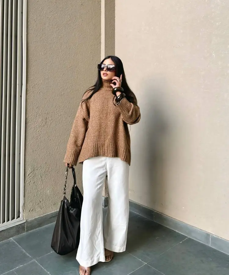 Uma mulher com cabelo liso e óculos de sol grandes veste um suéter oversized de tricô marrom e calças brancas largas. Ela segura uma grande bolsa preta e usa sandálias de salto. O fundo é uma parede neutra, com uma grade branca à esquerda.