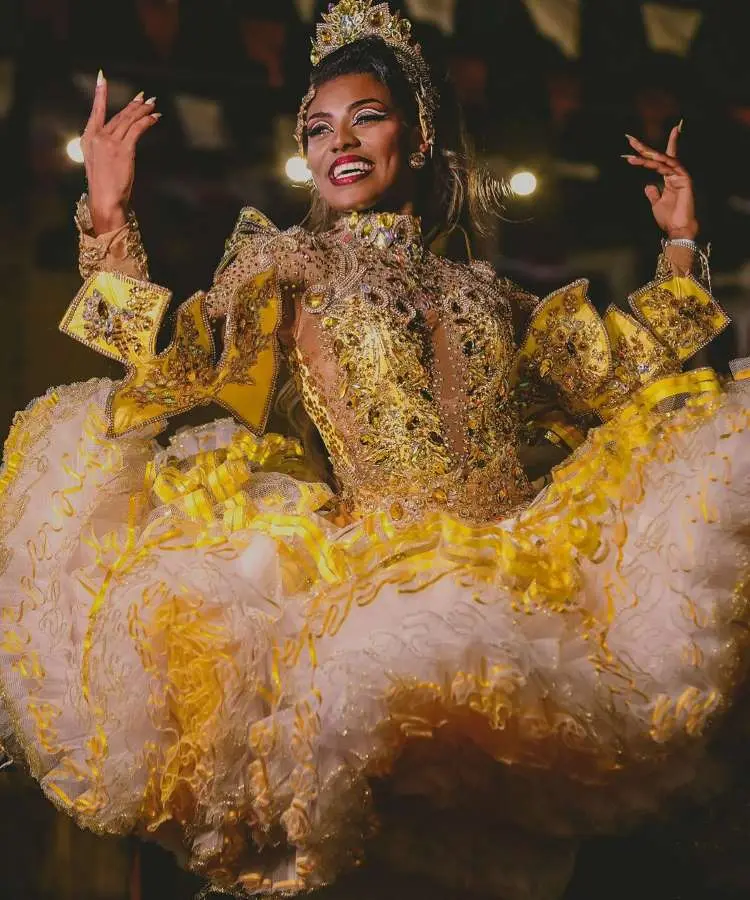 Imagem de uma rainha da quadrilha em festa junina, vestindo um traje dourado e branco com bordados e saia volumosa. Ela sorri e eleva os braços, demonstrando a vivacidade e a alegria das celebrações juninas, com luzes ao fundo.