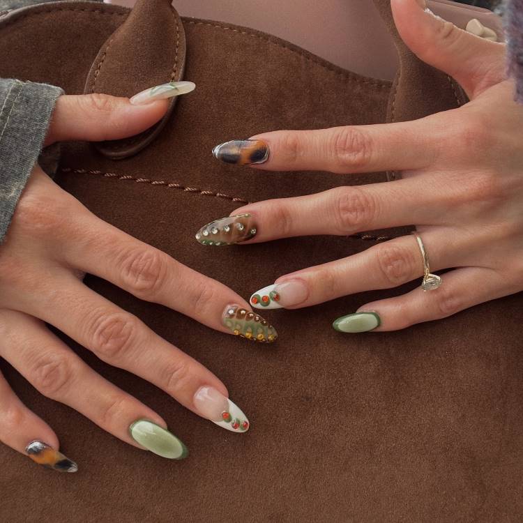 Unhas de gel decoradas com design variado, incluindo tons de verde, detalhes marmorizados, pedrarias 3D e esmaltes translúcidos. O visual sofisticado combina texturas e elementos criativos, destacando-se contra o fundo marrom de uma bolsa de couro, criando equilíbrio elegante.