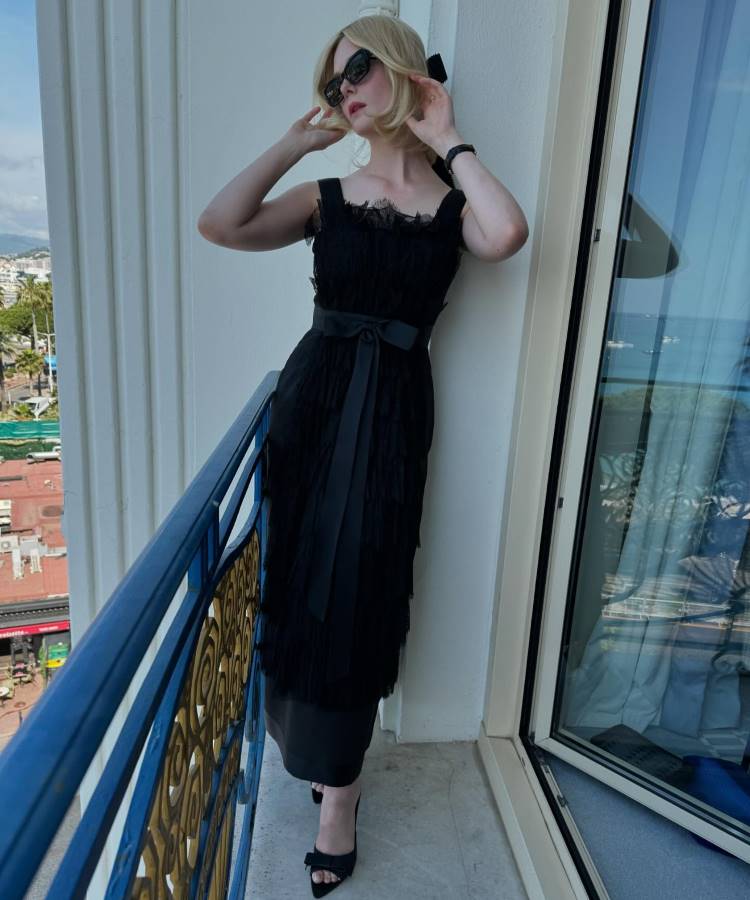 Uma mulher loira com óculos de sol escuros e um laço preto no cabelo posa em uma varanda. Ela veste um vestido preto midi sem mangas com detalhes em renda e um cinto de cetim. Sapatos peep-toe pretos com laços completam o visual.