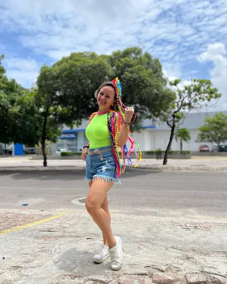 Foto de mulher de pele clara com body verde, shorts jeans e ombreira de fitas coloridas