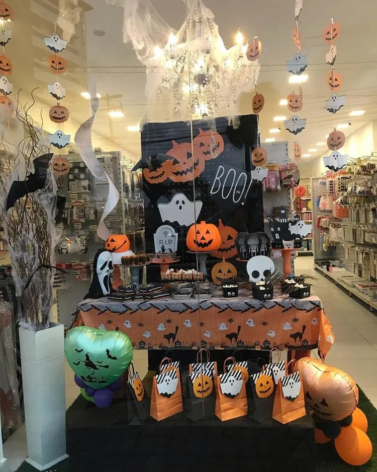Vitrine de Halloween com mesa com painel preto de abóboras e fantasmas, fios pendurados com desenho de abóboras e fantasmas, doces com embrulhos temáticos, desenhos de morcegos e sacolas de halloween 
