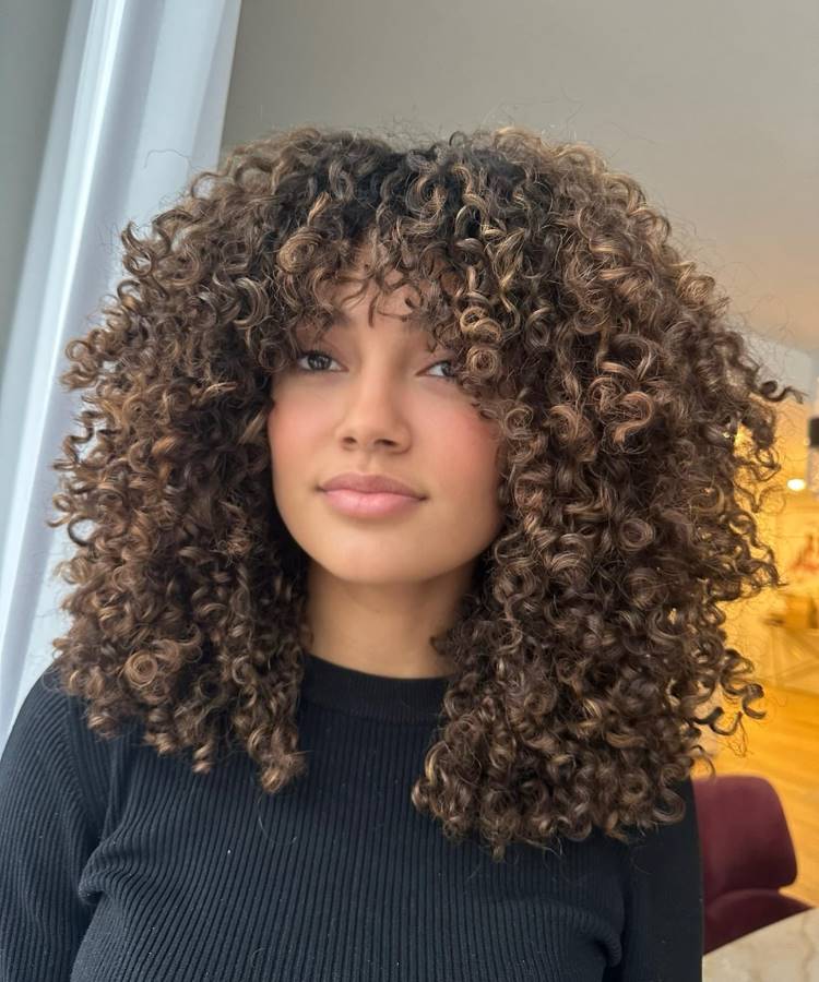 A imagem mostra uma jovem com cabelos cacheados volumosos, com mechas claras, e franja caindo sobre a testa. Ela veste uma blusa preta canelada e olha diretamente para a câmera com uma expressão calma. O fundo apresenta um ambiente interno com luz natural.







