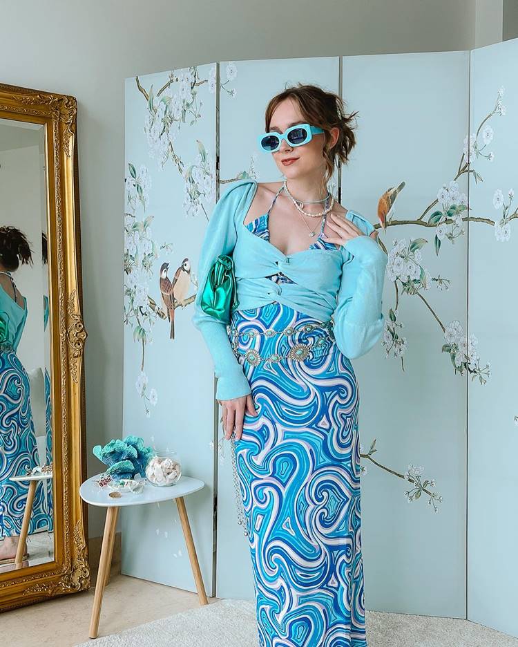 Menina com vestido Azul com listras em formato de coração top azul por cima bolsa verde e óculos azul
