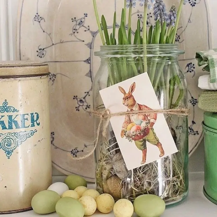 Pote de vidro decorado com gravura de coelho segurando ovos coloridos, preenchido com flores lilases e musgo seco. Ao lado, lata vintage bege, ovos de Páscoa em tons pastéis e toalha verde. Fundo com louça decorativa retrô.